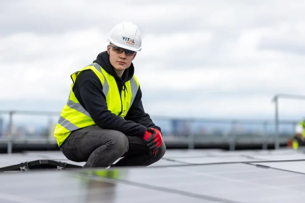 A man wearing a hard hat and safety vest kneels on a solar panel, representing the emerging field of green jobs in sustainability.
